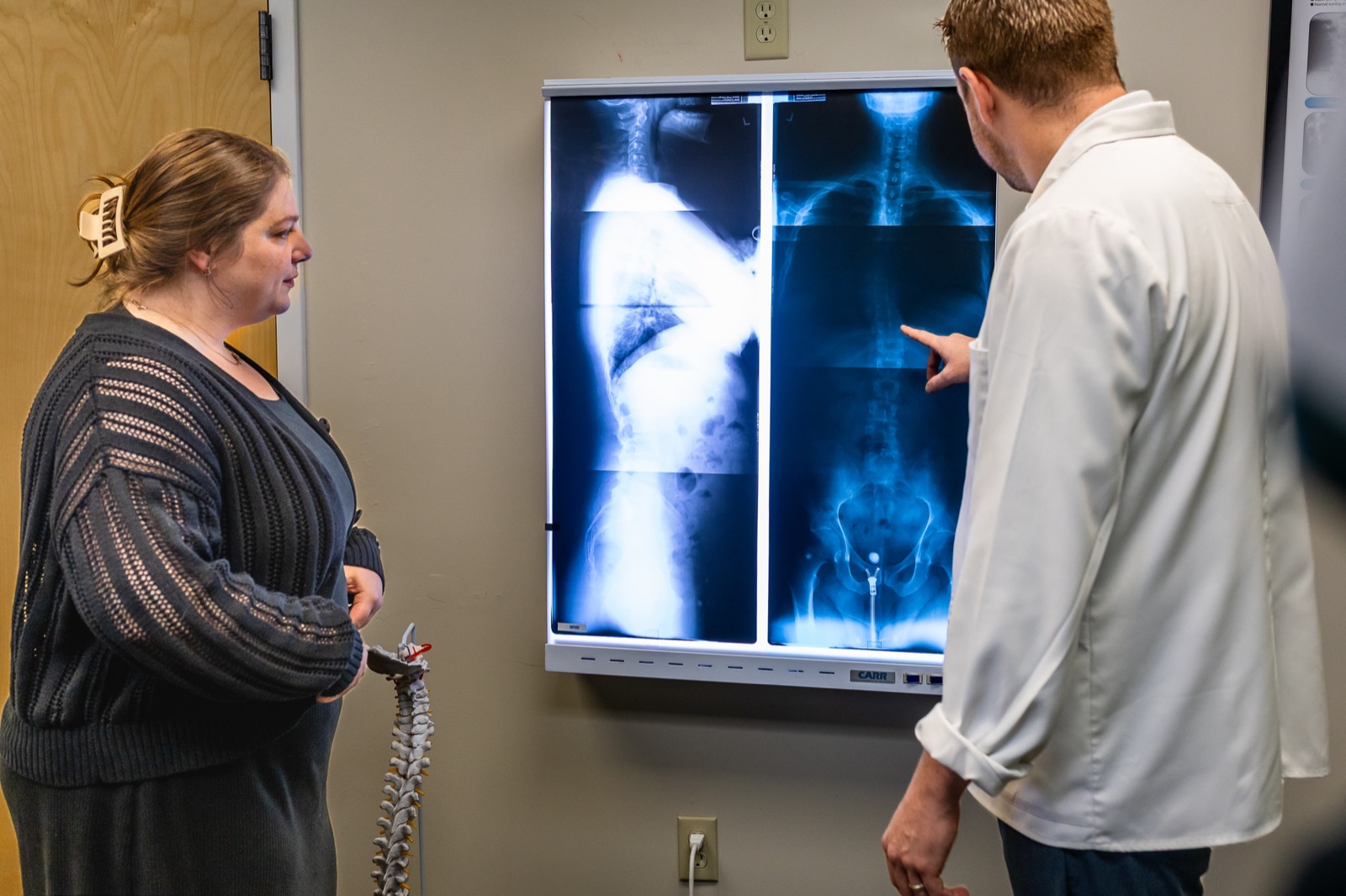 Dr. Connor Knapp pointing at findings on a lit full-spine X-ray with a patient